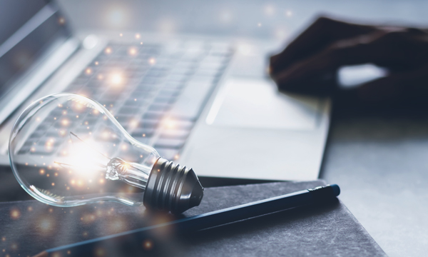A stock photo of a lit up lightbulb laying on its side on top of a notebook with a pencil. A laptop with a person's hand resting on the track pad is in the background.