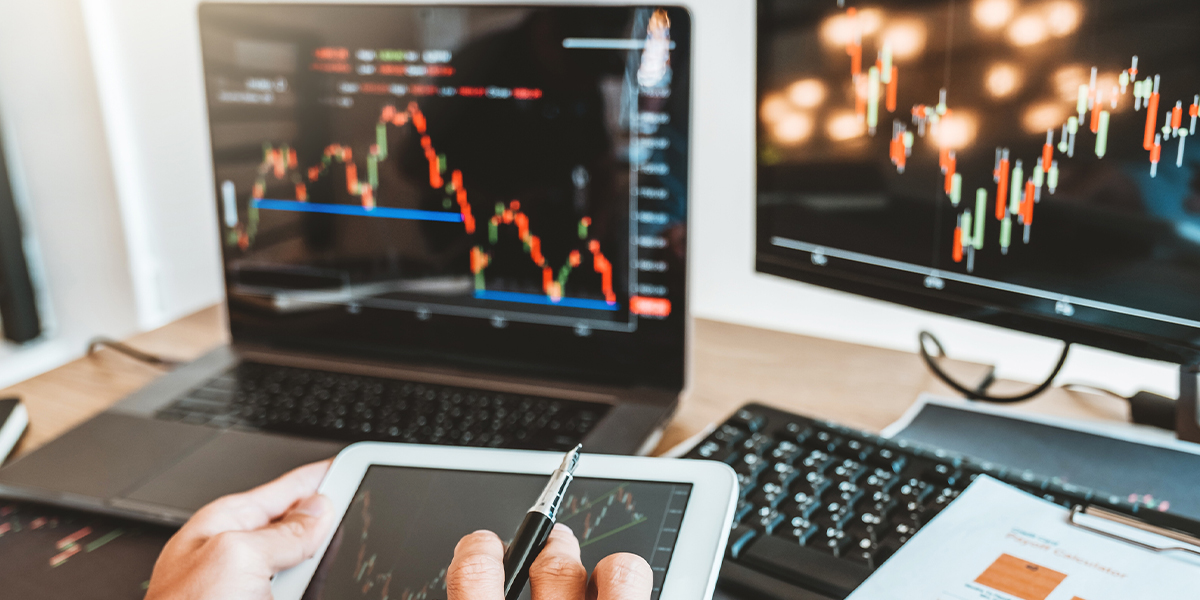 A stock image of a laptop and a computer monitor showing a stock market chart and hands holding a tablet also showing a stock market chart